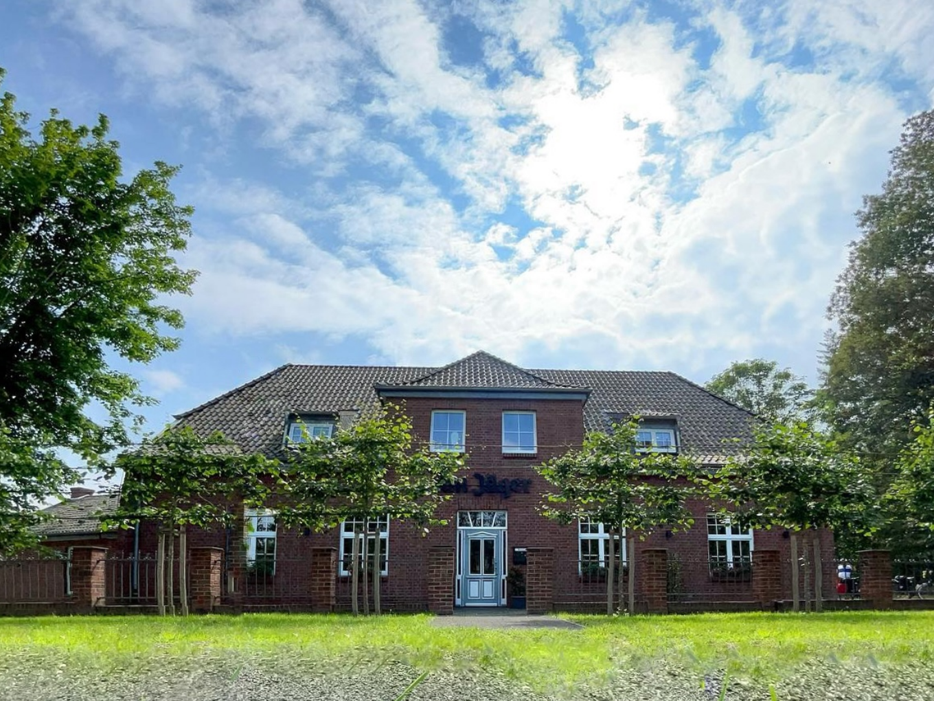 Backsteinfassade des Restaurants Am Jäger mit weißer Eingangstür, grüner Wiese und Bäumen unter blauem Himmel.