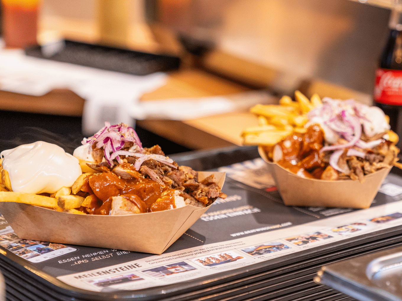 Zwei Portionen Döner mit Pommes, Zwiebeln und Sauce in Pappschalen auf einem Tisch mit Speisekarte