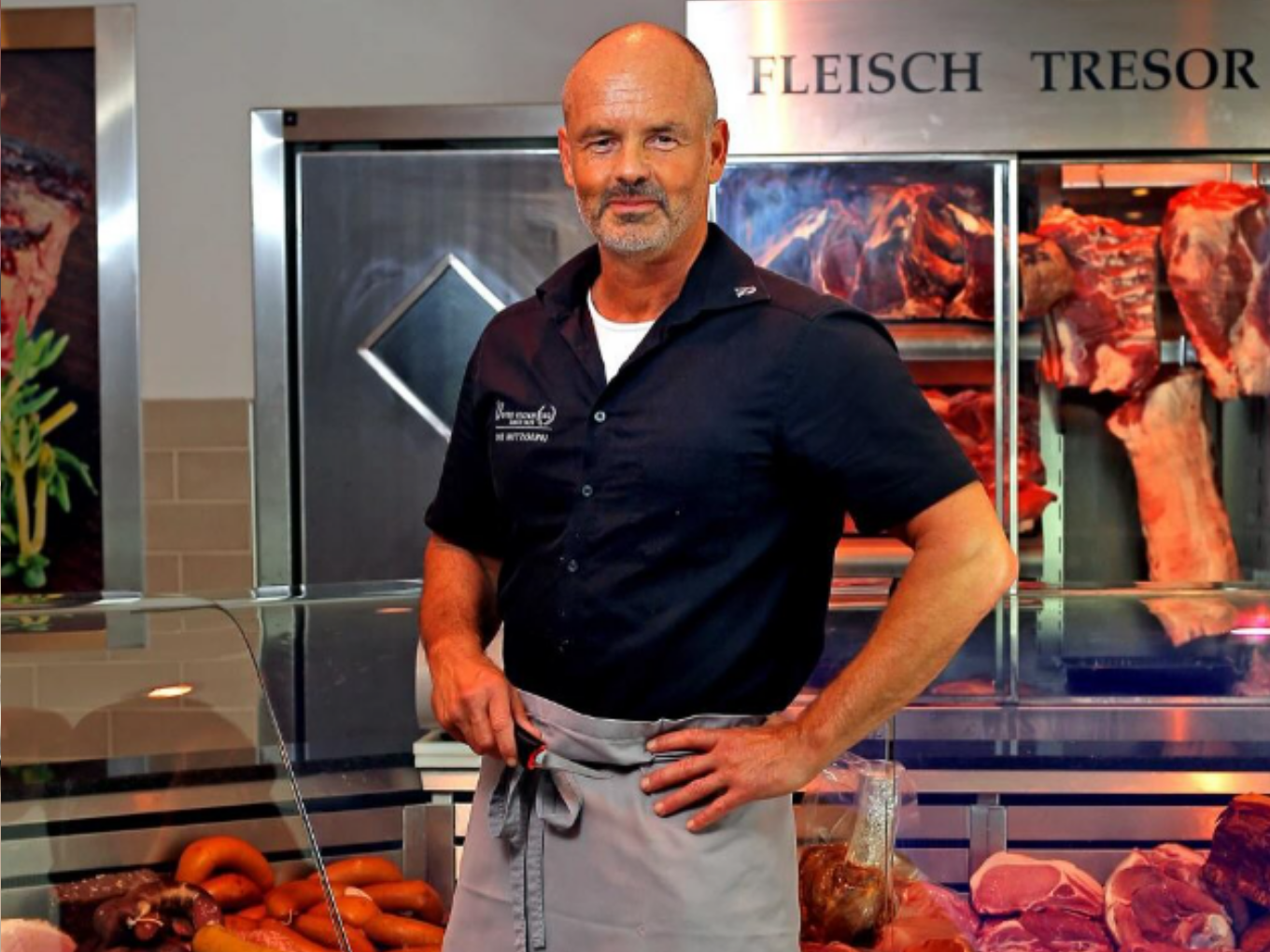Ein Metzger steht mit in den Hüften gestemmten Armen vor einer Fleischtheke, im Hintergrund Fleischstücke im „Fleisch-Tresor“.