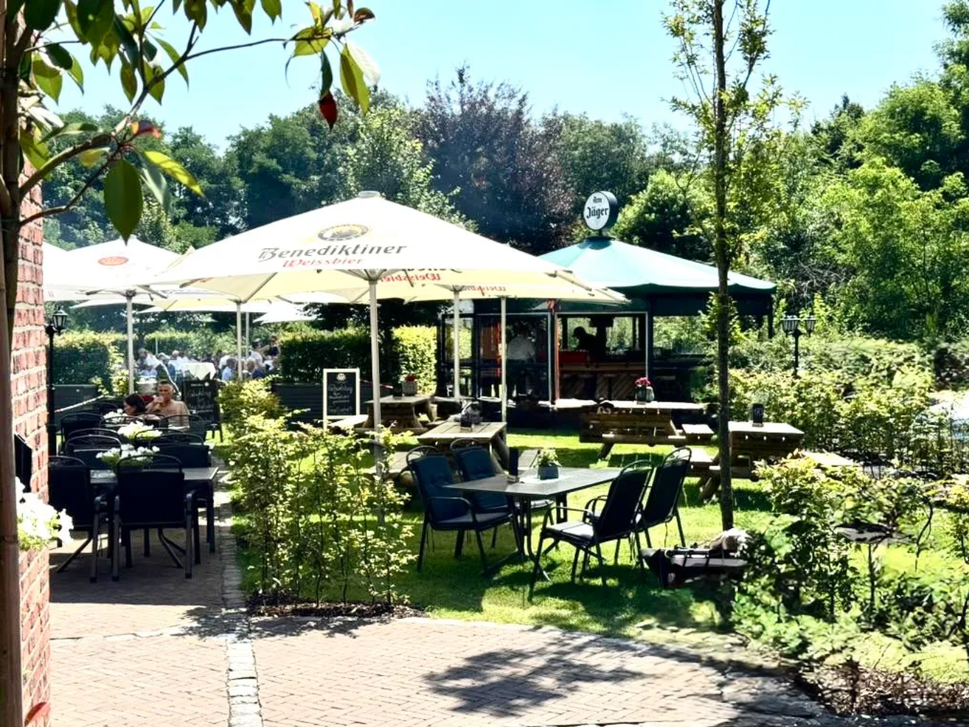 Sonniger Biergarten des Restaurants Am Jäger mit Tischen, Sonnenschirmen und Sitzplätzen im Grünen.