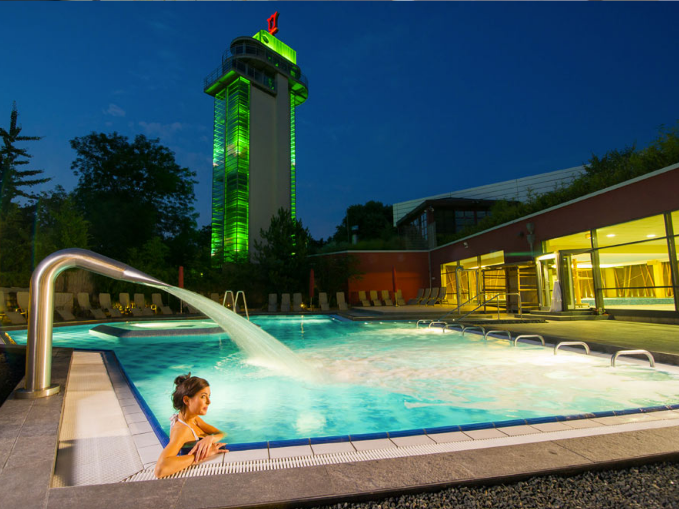 Beleuchtetes Außenbecken der Grugapark-Therme am Abend mit Wasserfontäne und grün angestrahltem Turm.