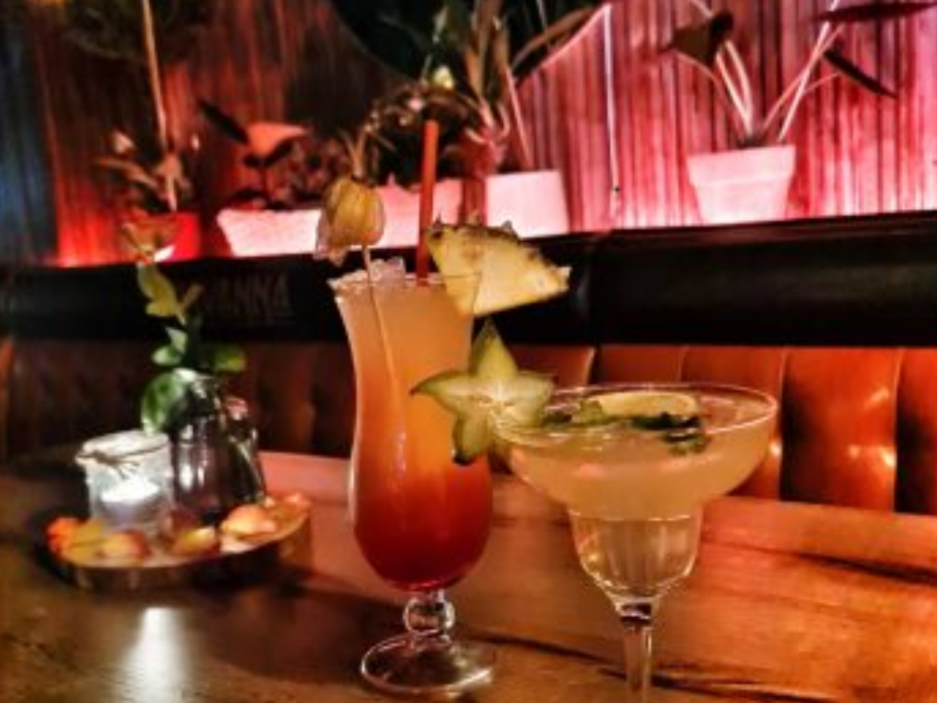 Zwei bunte Cocktails auf einem Holztisch, dekoriert mit Sternfrucht und Limette, im Hintergrund Pflanzen und eine gemütliche Sitzbank mit roter Beleuchtung.