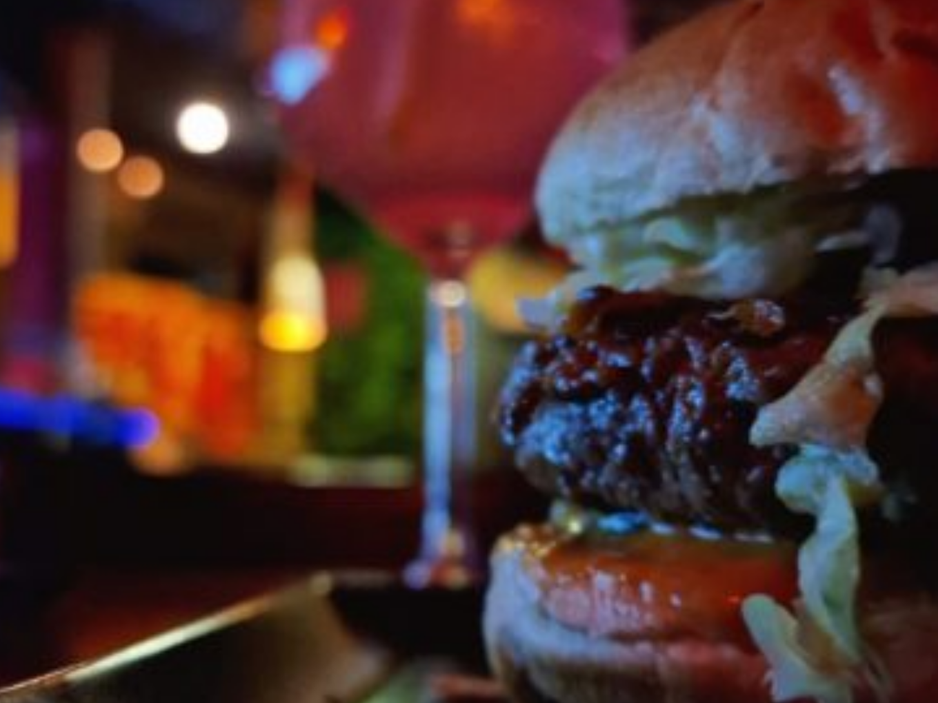 Ein saftiger Burger mit Salat und Tomate im Vordergrund, im Hintergrund unscharfe Lichter und ein Cocktailglas in einer stimmungsvoll beleuchteten Bar.