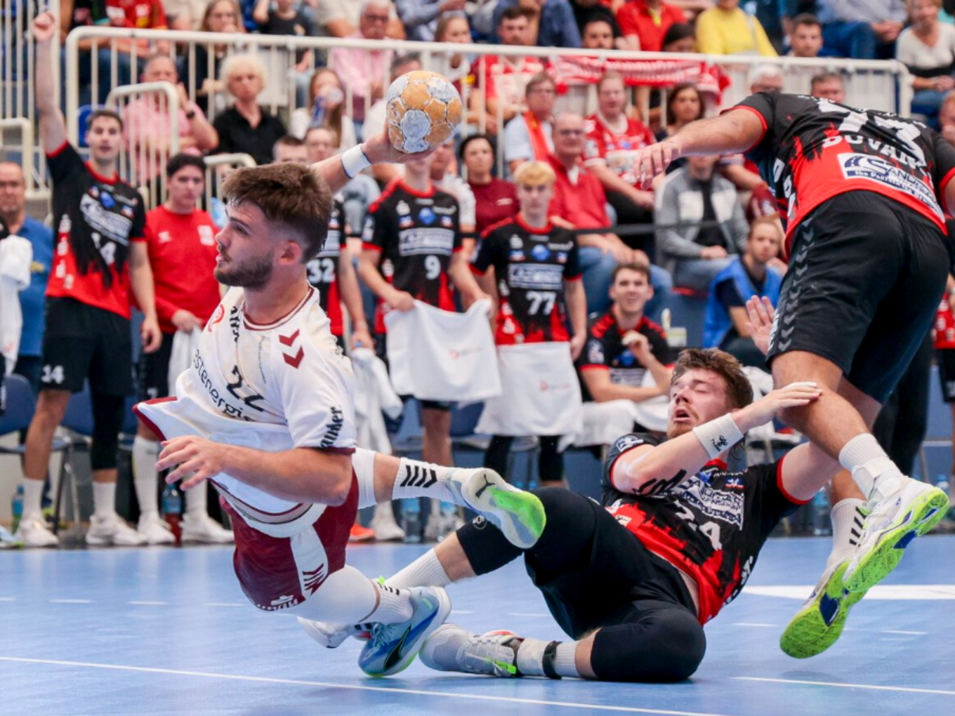 Handballspieler im weißen Trikot beim Sprungwurf, bedrängt von zwei Gegenspielern in einer vollen Halle