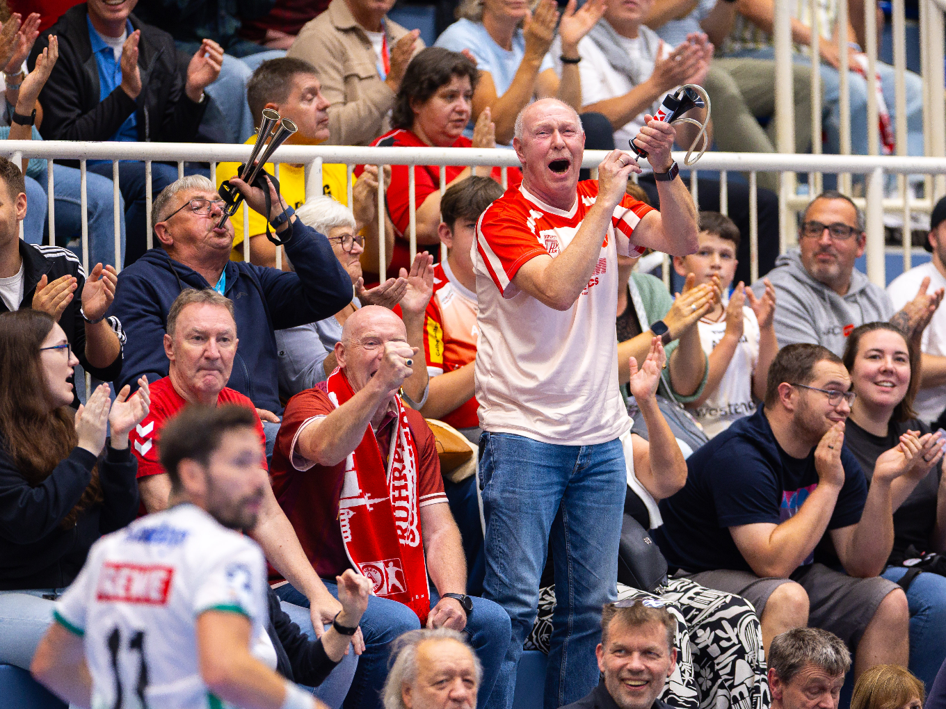 Begeisterte Fans in roten Trikots jubeln bei einem Handballspiel, einige blasen Vuvuzelas und klatschen.