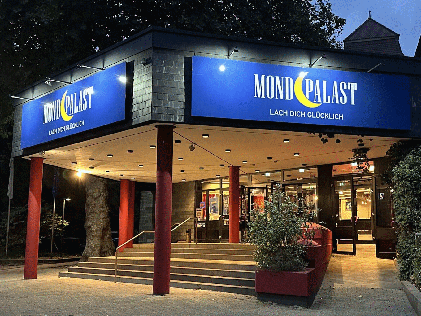 Das Bild zeigt den Haupteingang des Mondpalastes bei Abenddämmerung. Über dem Eingang befindet sich ein großes, blaues Schild mit der Aufschrift "Mondpalast" und dem Slogan "Lach dich glücklich".