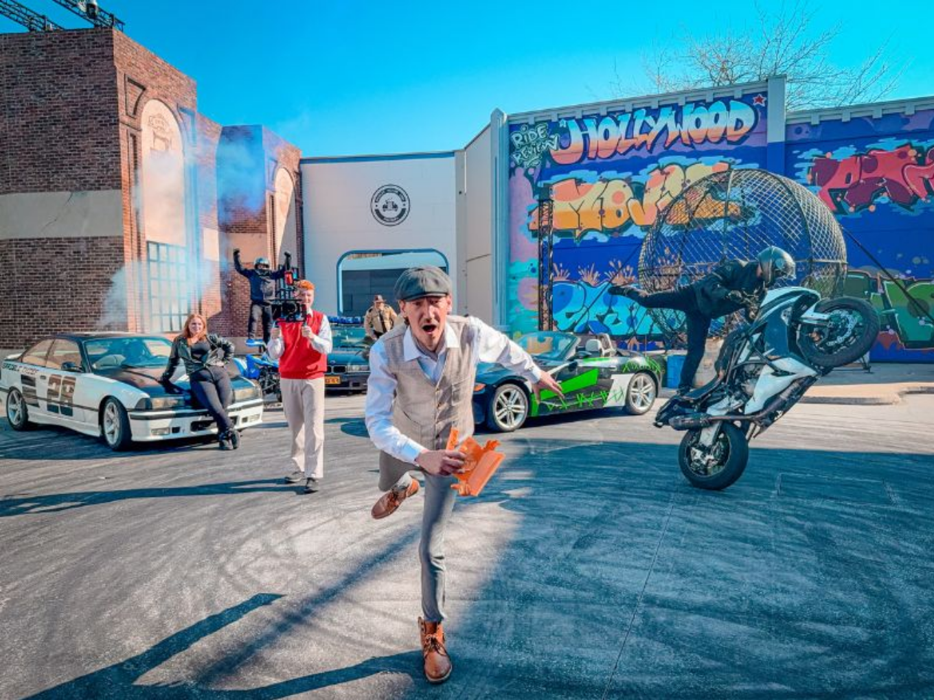 Actionreiche Szene mit Autos, Motorrad-Stunt und Menschen vor einer Kulisse mit Graffiti und Filmstudio-Atmosphäre.
