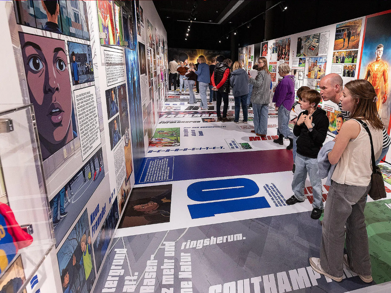 Besucher betrachten eine Wand mit Comic-Illustrationen und Texten in einer Ausstellung.