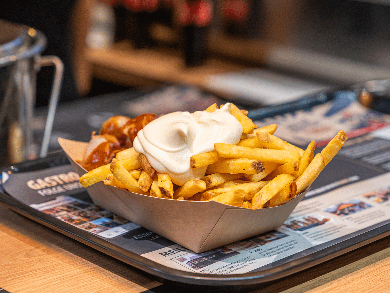 Portion Pommes mit Mayo und Currywurst in einer Pappschale auf einem Tisch mit Speisekarte