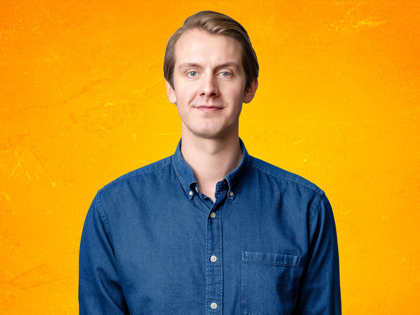 Comedian Jonas Greiner im blauen Hemd vor orangefarbenem Hintergrund