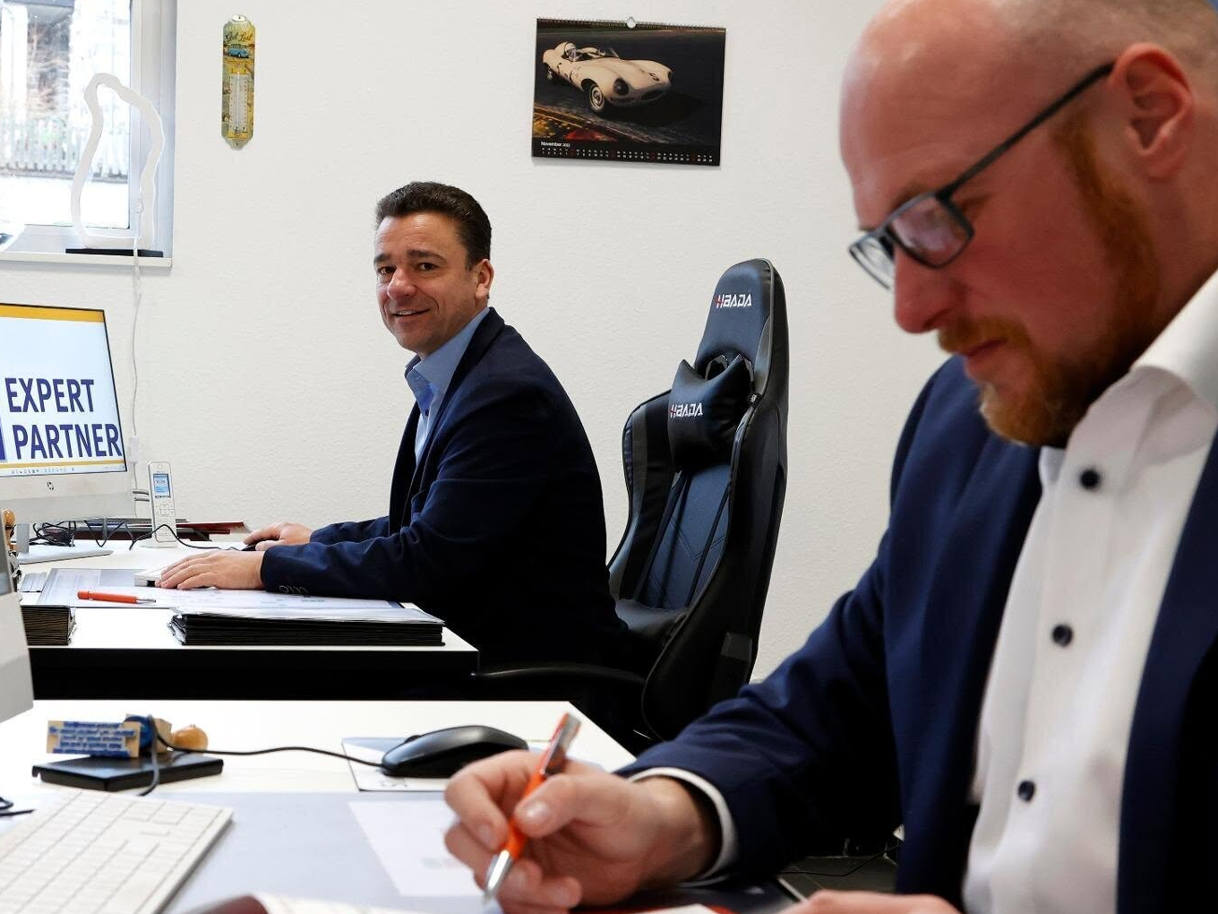 Zwei Männer im Büro, einer arbeitet am Computer, der andere schreibt etwas auf.