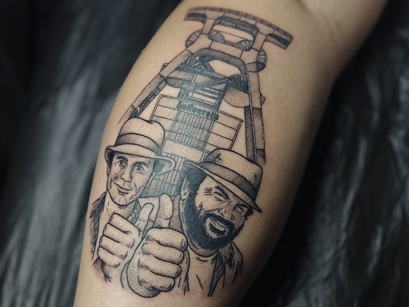 Fineline-Tattoo zweier Männer vor einem Förderkorb mit erhobenen Daumen