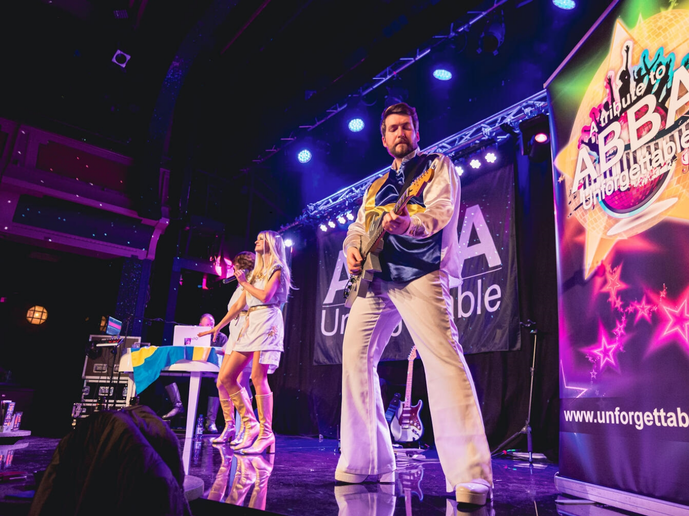 Drei Musiker auf einer Bühne bei einer ABBA-Tribute-Show. Ein Mann im Vordergrund spielt E-Gitarre und trägt ein blau-weißes Bühnenkostüm. Zwei Frauen im Hintergrund singen, eine davon steht an einem Keyboard