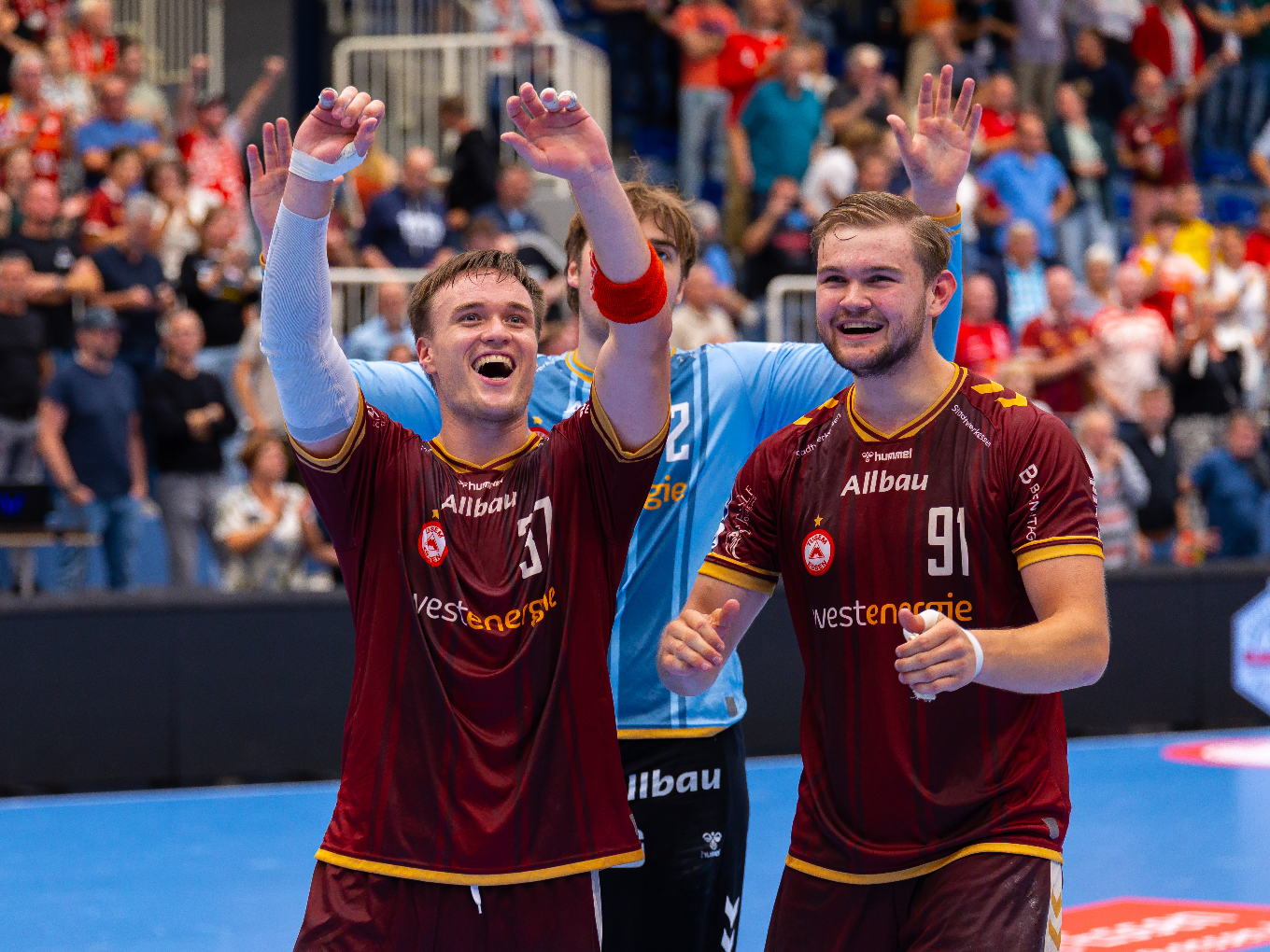 Drei Handballspieler von TUSEM Essen feiern nach dem Spiel mit erhobenen Armen vor jubelnden Fans in der Halle