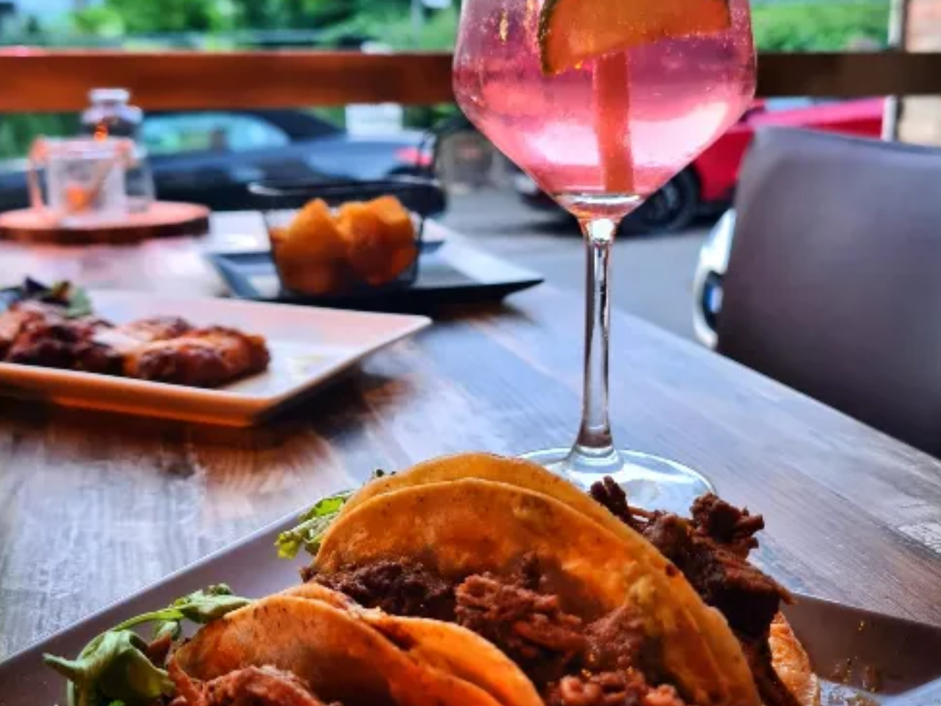 Drei Tacos mit Fleischfüllung auf einem Teller, daneben ein Glas mit pinkem Cocktail und Orangenscheibe, im Hintergrund weitere Speisen und ein Fenster mit Blick nach draußen.