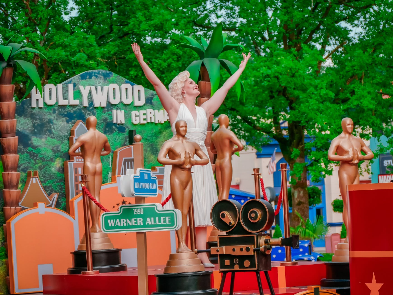 Eine Frau im weißen Kleid steht mit ausgebreiteten Armen auf einer Bühne, umgeben von Oscar-Statuen und Palmen, im Hintergrund steht „Hollywood in Germany“.
