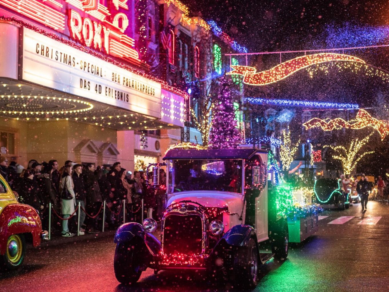 Oldtimer-Auto mit bunten Lichtern fährt durch eine festlich beleuchtete Straße, im Hintergrund viele Zuschauer.